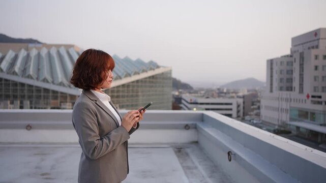 夕暮れのビル屋上でスマートフォンを操作し物思いにふける若い会社員女性