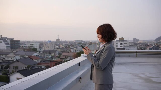 夕暮れのビル屋上でスマートフォンを操作し物思いにふける若い会社員女性