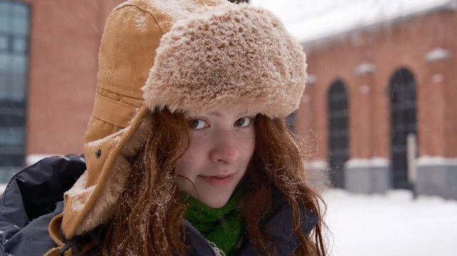 Cinematic close up portrait of a teenage girl with red hair wearing a warm ushanka hat, against the backdrop of an old city during gentle snowfall. snowflakes to fall softly