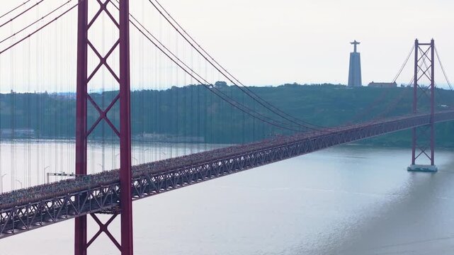 Cinematic aerial drone shot of thousands of long-distance runners crossing the iconic red 25th April suspension bridge in Lisbon, Portugal, with the Cristo Rei monument visible. Dark grade