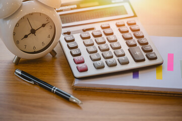 Calculator and alarm clock notebook with pen on wood table with blurred bokeh background...
