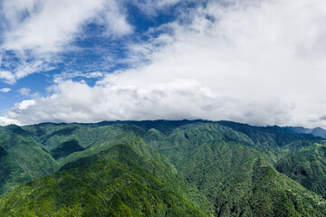 Naklejka premium Majestic green mountain range covered with dense forest under a bright blue sky with white clouds.