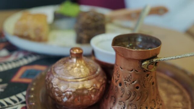 Ornate copper cezve and matching sugar bowl on decorative metal tray, placed on top of traditional bosnian patterned textile. Confectionery and ceramic cup at background. Traditional bosnian coffee