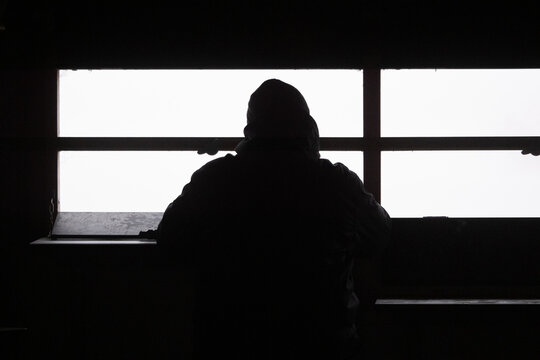 Silhouette of an unidentified person standing and looking out through a bright window from a dark indoor space. The strong contrast between light and shadow creates a dramatic and emotional atmosphere