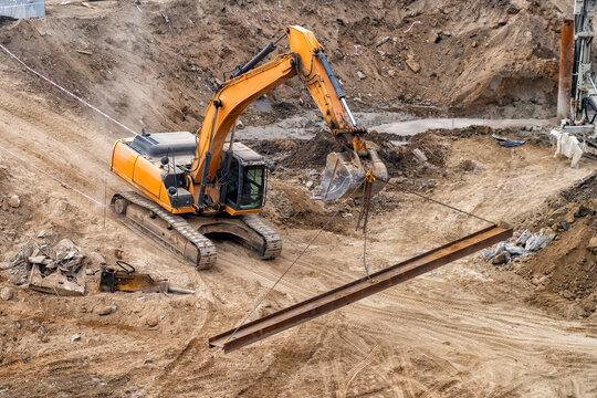Heavy yellow excavator lifting long steel beam using chains in deep construction foundation pit surrounded by excavated brown earth and dirt