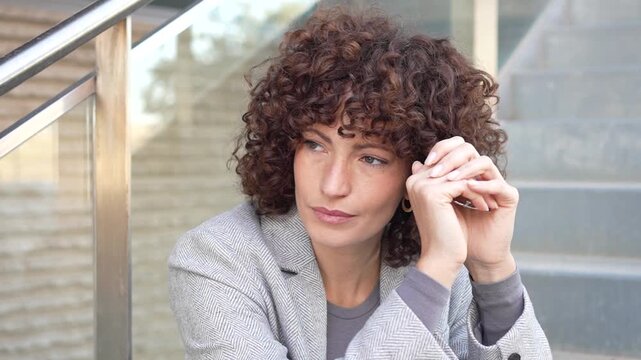 A thoughtful woman, with beautiful curly hair, is sitting on the stairs in a contemplative manner