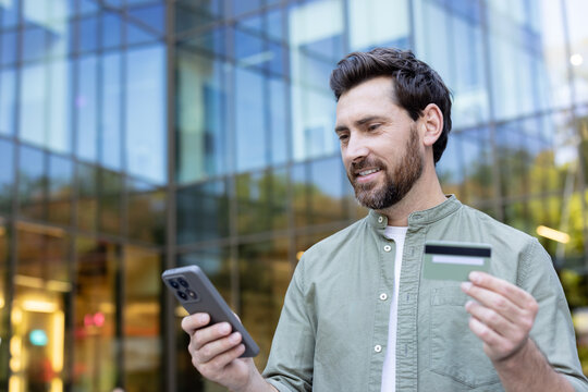 Man holding a smartphone and a credit card, smiling while making an online purchase or banking transaction outdoors in a city setting, reflecting modern digital commerce and financial technology - Powered by Adobe
