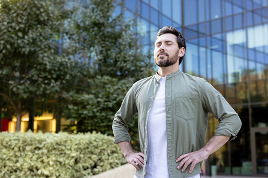 Man standing with eyes closed and hands on hips, taking a deep breath of fresh air and finding a calm, mindful moment of relaxation in an urban business district