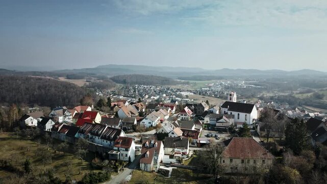 Striking forward drone flight over Aach village rooftops, church steeples, winding paths, autumn foliage, meadows, and surrounding wooded hills. Perfect for travel docs, regional ads