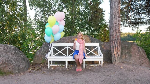Young woman sit on bench with candy, look straight to camera, bunch of balloons float in air beside. Nature environment, large stones and tree trunk, green leaves on background