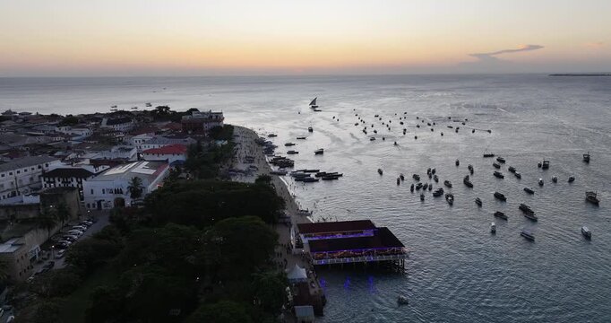 Cinematic drone shot of sunset over the night market in Stone Town, Zanzibar, with traditional boats along the waterfront. Warm colors, coastal atmosphere, and lively evening vibes create a perfect tr