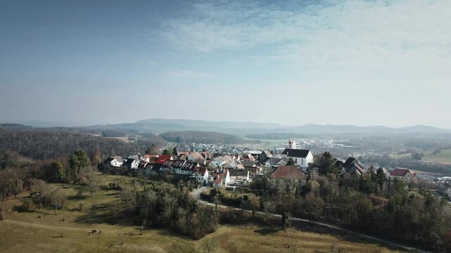 Compelling forward drone glide toward Aach village cluster with church tower, autumnal trees, grassy meadows, and encircling hills in hazy sunlight. Perfect for travel docs, regional ads