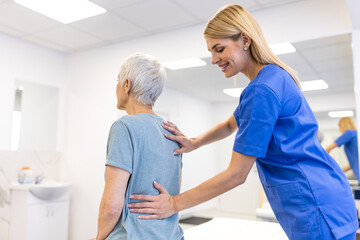 Female physical therapist in blue scrubs providing postural correction and back support to an elderly woman in a rehabilitation center, helping with spinal alignment and geriatric healthcare.