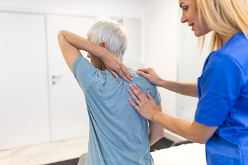 Female physical therapist in blue uniform guiding a senior woman through shoulder and upper back stretches during a rehabilitation session in a modern clinic to treat muscle tension and stiffness.