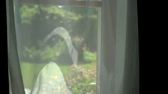 White sheer curtains gently billow in the wind over a window, revealing a beautiful, blurred green garden with colorful flowers outside. Serene and fresh atmosphere.