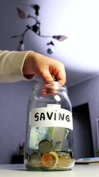 Hand dropping coins into glass savings jar on table. Close up of fingers inserting money into transparent container, representing simple financial planning and goal achievement in everyday life