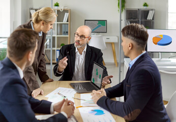 Business team meeting with manager leading discussion. Colleagues in a bright office review planning charts on a laptop and align goals. Teamwork and corporate strategy planning concept.