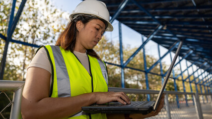 Fototapeta premium A person is operating a laptop at a construction site under a metal structure. The individual is dressed in a safety vest and hard hat. Trees and clear sky are in the background