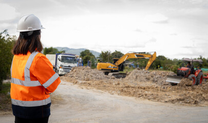 A person wearing a hard hat and safety jacket stands by the road watching workers perform tasks...