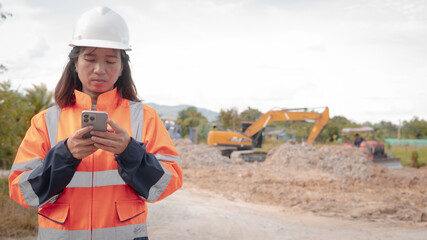 A construction worker in safety gear looks at her phone on a work site. Heavy machinery is active in the background, moving dirt and rocks