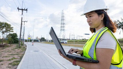 A construction worker in a hard hat and safety vest stands by the road using a laptop. Power lines run in the background on a sunny day © Happy Photo