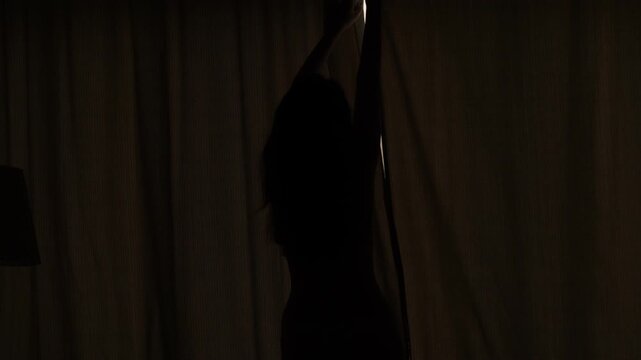 Half-length silhouette of young adult woman against window, closing curtains at early morning time. Day break and dim first light outside, girl shut blackout fabric and room become dark