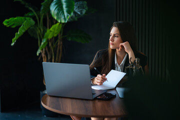 A thoughtful businesswoman sits at a desk, contemplating her work with a laptop and documents, surrounded by greenery in a modern office.