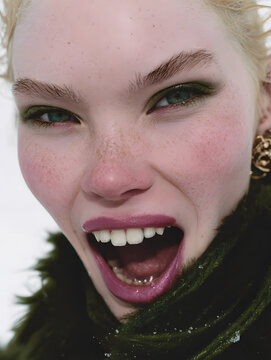 A smiling blonde lady in the snow, wearing a green coat and scarf, against a snowy backdrop, bathed in sunlight. 