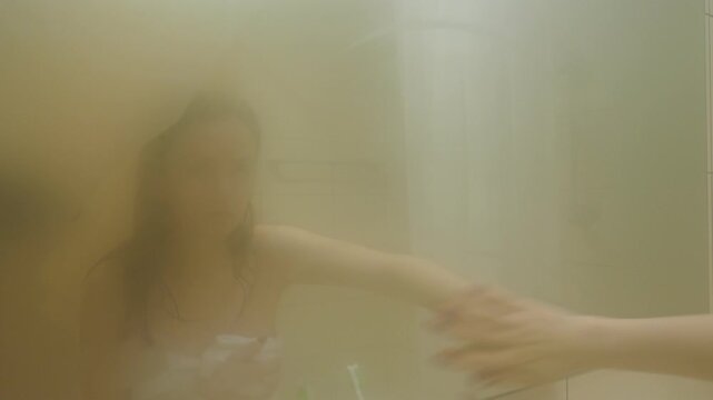 Young adult woman wipe mirror glass in foggy bathroom, look to hazy reflection after bathing. Thick vapory air in bath-room after taking hot shower. Woman stay wrapped into white towel