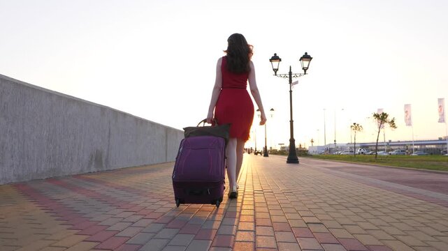 Woman walk with wheeled bag towards evening sun, slow motion shot, dolly camera follow behind. Nice empty promenade at sea front, modern embankment at Adler city. Concrete wall on left