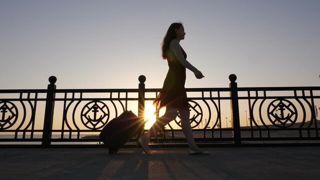 Beautiful young woman walk with trolley case at quay, sun flick through fencing, silhouette shot. Girl pull wheeled suitcase, come at nice embankment near Black Sea, smooth low angle tracking shot