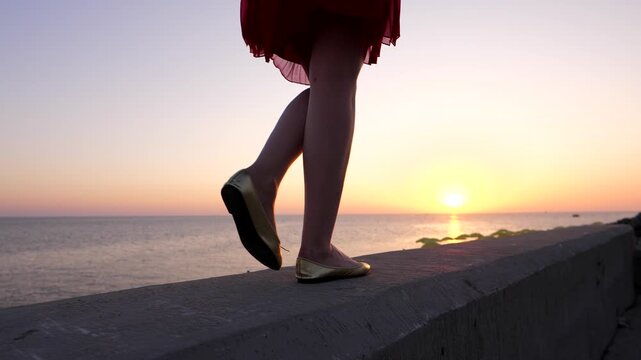 Woman legs pace at embankment parapet against sunset sky and calm sea, slow motion dolly shot. Girl enjoy beautiful evening time at waterfront. Warm breeze from calm water, golden sun move down