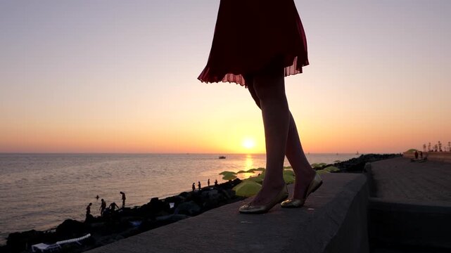 Woman legs stand on parapet at sea embankment, light summer dress skirt wave on breeze. Clear sunset sky, low sun shine on background. Dark beach, some tourists resting around area.