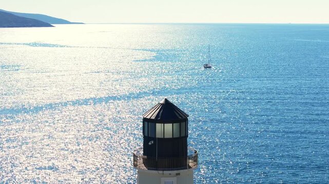 Aerial view frames a small octagonal lighthouse lantern and balcony, a vast sunlit sea, rocky headlands left, and a sailboat drifting right at midday.