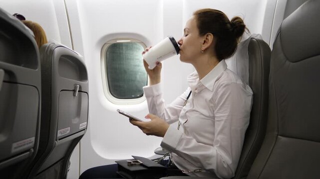 Tired business woman drink coffee from large paper cup, sitting in airliner before departure. She hold smarphone in hand, do not interrupt from work on go. Typical busy lifestyle, no time to rest