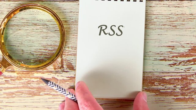 On a distressed wood table, a hand writes the letters RSS on a notepad. A golden magnifying lens and a patterned pen are positioned nearby