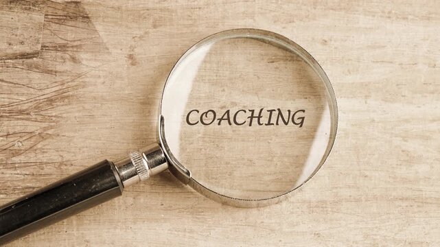 A magnifying glass slowly reveals the word COACHING written on a textured surface, transforming from abstract marks to a clear concept. Exploration and discovery