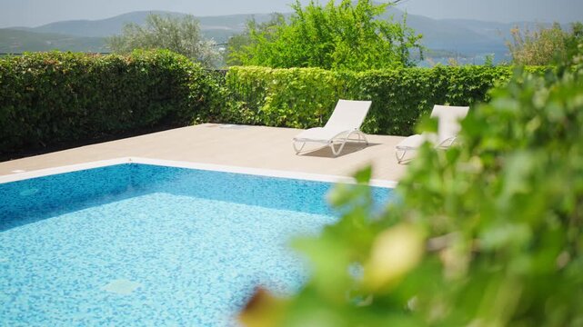 A private outdoor pool with blue mosaic tiles and two white loungers on a beige deck, ivy hedges, soft focus leaves, rolling hills, and a gentle pan in warm summer light.