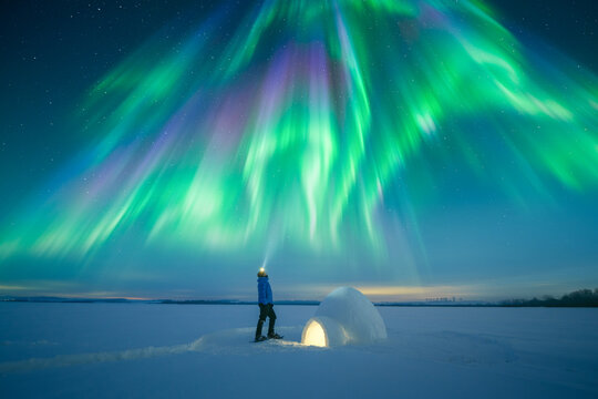 Man with flashlight near snow igloo lighted from the inside under incredible starry sky with aurora borealis, winter night landscape, polar lights, northern lights