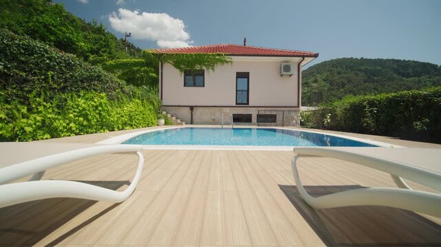 Low angle view of a private villa, red tiled roof, AC unit, blue mosaic pool and white lounge chairs as the camera dollies forward, wide and sunlit at midday