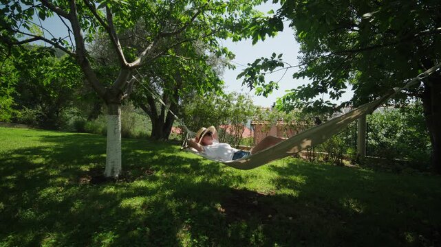 A person reclines in a canvas hammock between leafy trees as it sways, in a sun dappled residential garden with terracotta roof houses visible beyond a low hedge.