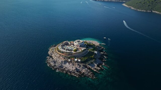 Aerial view of Mamula island in the Adriatic shows concentric fortress, terraces, pool and helipad, with speedboats carving wakes amid turquoise shallows and mountains.