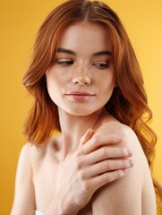 A woman with red hair and freckles gently touches her shoulder against a bright yellow background. Beauty and skincare concept.