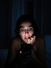 A woman with wide eyes and a fearful expression stares at her phone in a dimly lit room. Emotional reaction to digital content.