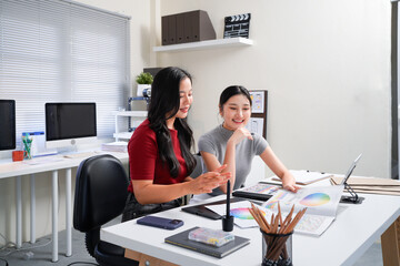 Two professional women collaborating on creative design project, discussing colorful color swatches, hues, and palettes at modern office workspace table. Ideal for wallpaper, poster