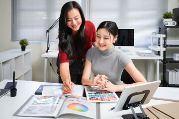 Two professional women collaborating on creative design project, discussing colorful color swatches, hues, and palettes at modern office workspace table. Ideal for wallpaper, poster