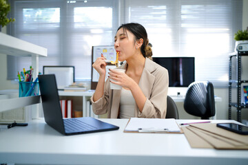 woman eating instant noodles sitting in office with laptop. Hungry female employee is having lunch at workplace