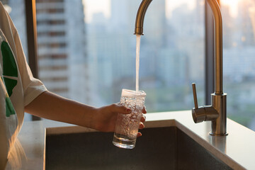 Girl fills a glass with clean filtered tap water for drinking in the kitchen at home. Window in background. Modern faucet and sink in home kitchen. © GALINA