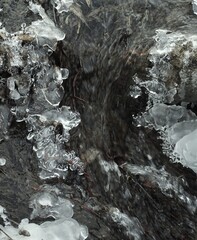 Woda w strumieniu przepływa przez lodowe szczeliny, otoczona zimowym krajobrazem. Lodowe formacje tworzą efektowny, zimowy wzór. © Adam