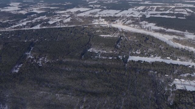 Aerial pan and descent show snow dusted pines near Nowy Lubiel, a frozen meandering river, striped canopy bands, rural cottages, and distant fields in winter light.